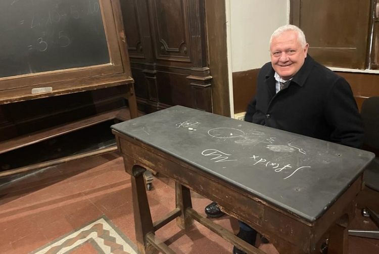 Seduto alla cattedra di un'aula storica, accanto alla lavagna